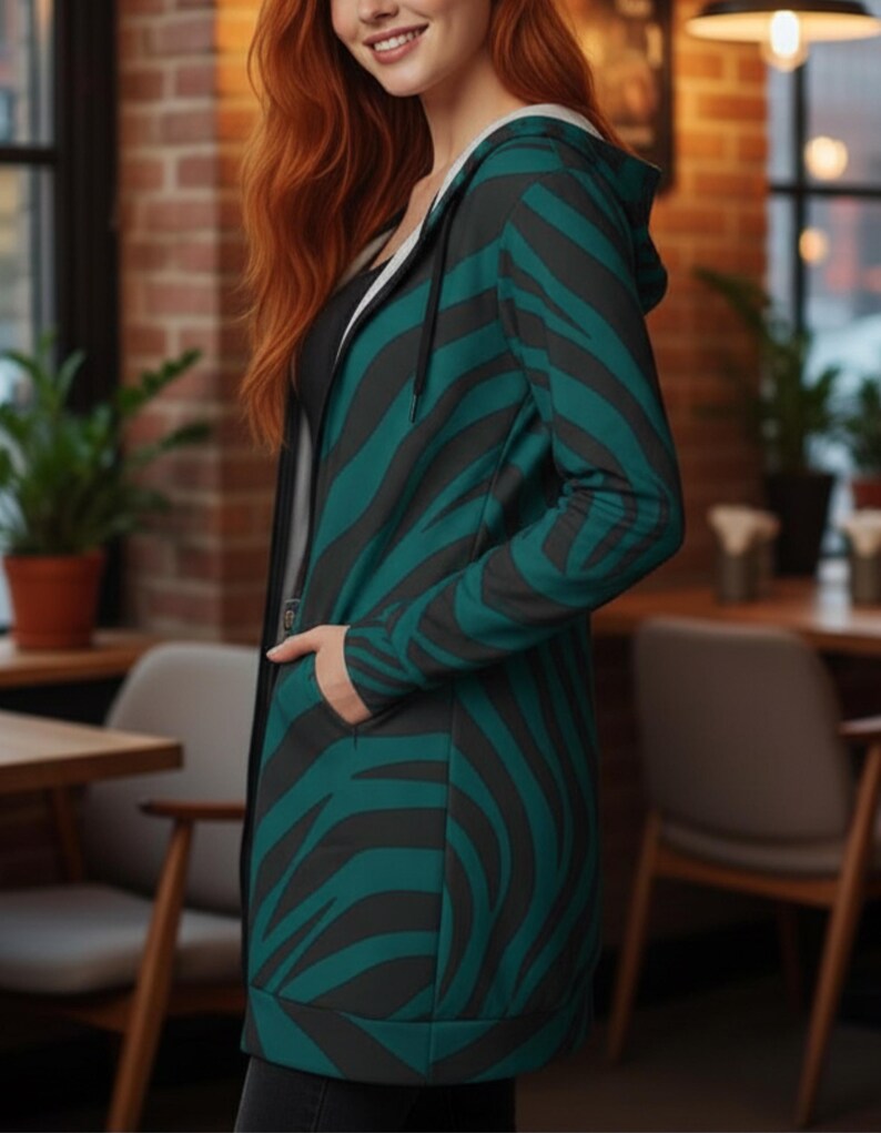 Peut inclure: Une longue veste &agrave; capuche avec un motif z&eacute;br&eacute; teal et noir. La veste a une fermeture &eacute;clair et des poches. L'int&eacute;rieur de la capuche est blanc. La personne porte un haut noir en dessous.