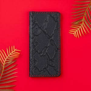 May include: A black phone case with a textured, cracked pattern and red stitching. The case is rectangular and appears to be made of leather. The background is a solid red color, with gold-colored palm leaves in the corners.
