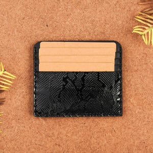Könnte beinhalten: Ein schwarzer Kartenhalter mit Schlangenmuster, der mehrere beige Karten enthält. Der Kartenhalter besteht aus einem glänzenden, strukturierten Material. Das Design ist schlicht und elegant, für Kredit- oder Visitenkarten.