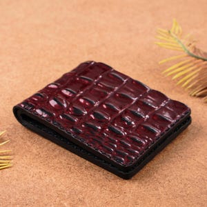 May include: A burgundy leather wallet with a crocodile skin pattern. The wallet is rectangular and has a black border. The texture of the leather is visible, and the wallet appears to be new.
