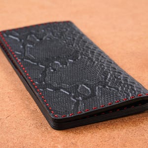 Può includere: Un portafoglio nero con motivo a pelle di serpente e cuciture rosse. Il portafoglio è rettangolare e sembra essere realizzato in pelle o materiale simile. Le cuciture sono visibili lungo i bordi.