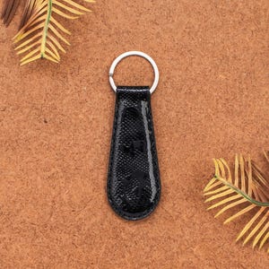 Op de afbeelding: Zwarte leren sleutelhanger met een zilveren sleutelring. De sleutelhanger heeft een getextureerd oppervlak en een glanzende afwerking. Het ontwerp heeft de vorm van een druppel. De achtergrond is een bruin oppervlak met gele en groene bladeren.