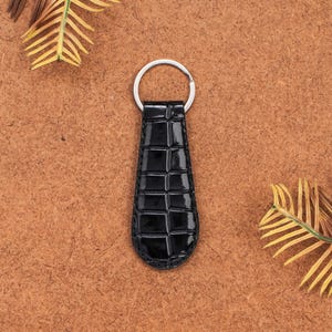 Può includere: Portachiavi in pelle nera con anello argentato. Il portachiavi presenta un motivo strutturato a forma di coccodrillo. Lo sfondo è di colore marrone caldo con alcune foglie secche.