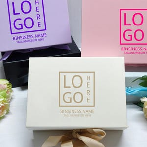Può includere: Tre scatole regalo rettangolari in lavanda, rosa e crema, ciascuna con un logo e testo. La scatola lavanda ha un logo viola, la scatola rosa ha un logo rosa e la scatola crema ha un logo dorato. Ogni scatola ha un nastro abbinato.