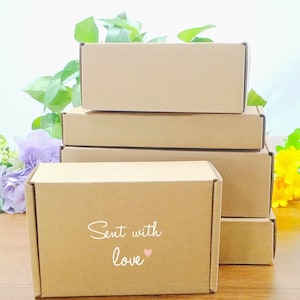 Op de afbeelding: Een stapel rechthoekige, bruine kartonnen dozen. Op de voorste doos staat in sierlijke letters "Sent with love", met een klein roze hartje. De dozen zijn netjes opgestapeld, wat suggereert dat ze klaar zijn voor verpakking en verzending.