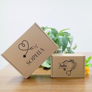 May include: Two brown cardboard boxes with black text and designs. One box has a stethoscope heart design and the name "SOPHIA". The other box has the name "Julie" with a floral design and stethoscope. The boxes are on a wooden surface.