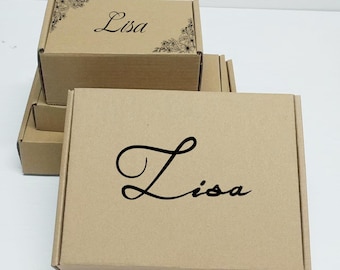 Op de afbeelding: Verschillende kleine, bruine kartonnen dozen gestapeld. De bovenste doos heeft de naam "Lisa" in zwart script, met bloemenhoekaccenten. De dozen zijn effen en hebben een eenvoudig, strak ontwerp, geschikt voor cadeaus of kleine items.