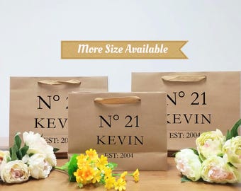 May include: Three brown paper shopping bags with handles, each printed with "N° 21 KEVIN EST: 2004" in black. The bags are arranged in front of a white background, with flowers in the foreground. A banner reads "More Size Available".