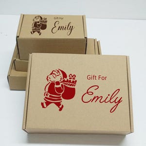 Peut inclure: Plusieurs boîtes cadeaux en carton marron empilées. Chaque boîte présente une illustration rouge du Père Noël portant un sac de cadeaux, avec l'inscription "Gift For Emily" en rouge.
