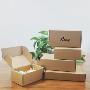 May include: A collection of brown cardboard boxes, some stacked and one open, revealing contents. The word "Rose" is written on one box. The boxes are on a wooden surface with green plants in the background.