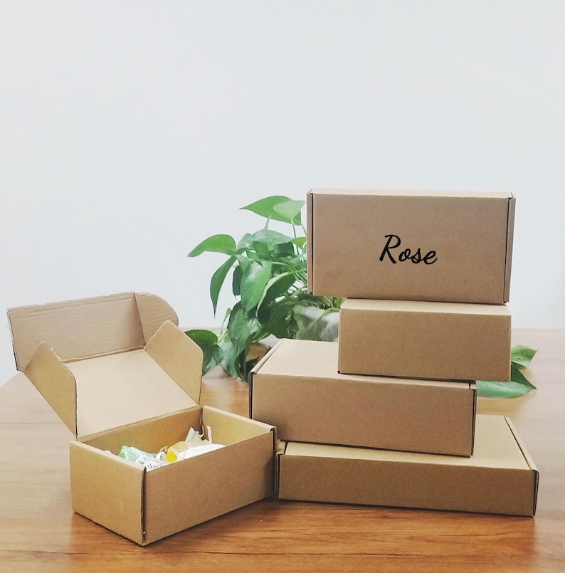 May include: A collection of brown cardboard boxes, some stacked and one open, revealing contents. The word "Rose" is written on one box. The boxes are on a wooden surface with green plants in the background.