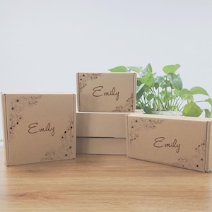 Puede incluir: Varias cajas de papel kraft con diseños florales en las esquinas y el nombre "Emily" impreso. Las cajas están dispuestas sobre una superficie de madera, con una planta en el fondo.