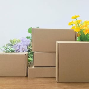 May include: Several brown cardboard boxes of varying sizes are arranged on a wooden surface. Artificial flowers in shades of yellow, green, and purple are placed near the boxes. The boxes are empty and ready for use.