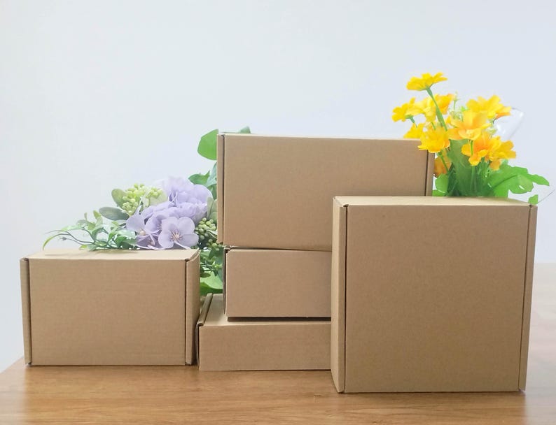 May include: Several brown cardboard boxes of varying sizes are arranged on a wooden surface. Artificial flowers in shades of yellow, green, and purple are placed near the boxes. The boxes are empty and ready for use.