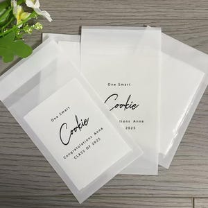 Może przedstawiać: Trzy białe papierowe torebki z czarno-białym wzorem. Torebki mają napis "One Smart Cookie" i "Congratulations Anna CLASS OF 2025".