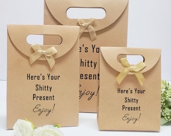 May include: Three brown paper gift bags of varying sizes. Each bag has a handle, a gold ribbon, and the text "Here's Your Shitty Present Enjoy!" printed in black. The bags are arranged on a white surface.