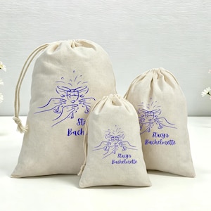 Puede incluir: Tres bolsas de cordón de color blanco roto de diferentes tamaños, cada una con un gráfico de líneas azules de manos brindando con copas de champán y el texto "Stacy's Bachelorette" impreso en azul en la parte frontal de cada bolsa.