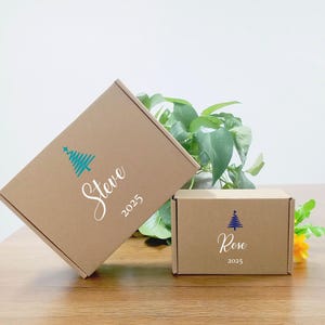 May include: Two brown cardboard boxes with teal Christmas tree designs and names. One box reads "Steve 2025" and the other reads "Rose 2025". The boxes are on a wooden surface with green foliage in the background.
