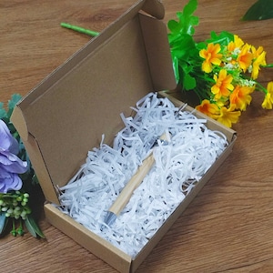 May include: A pen with a light brown wooden barrel and silver accents rests inside a cardboard box filled with white paper shreds. The box is open, revealing the pen, and is set on a wooden surface with yellow and purple flowers.