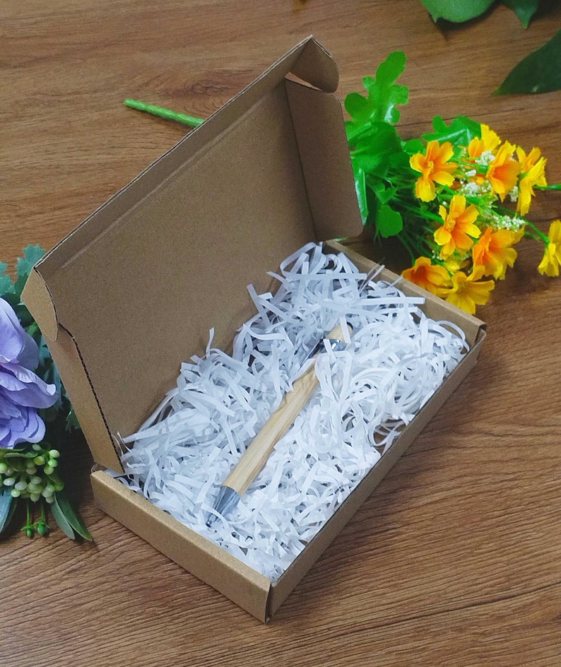 May include: A pen with a light brown wooden barrel and silver accents rests inside a cardboard box filled with white paper shreds. The box is open, revealing the pen, and is set on a wooden surface with yellow and purple flowers.