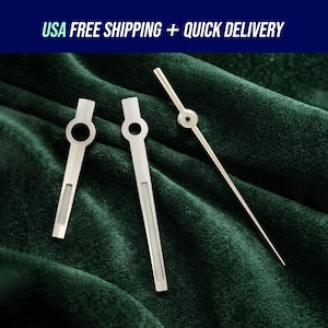 May include: Three silver watch hands on a dark green velvet surface. Two hands have a circular opening and a rectangular section. The third hand is long and thin with a circular detail at the end. Text at the top reads "USA FREE SHIPPING + QUICK DELIVERY".