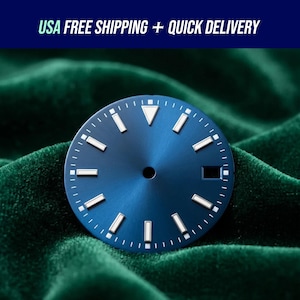 Pode incluir: Um mostrador de relógio azul com marcadores e ponteiros brancos. O mostrador tem um acabamento sunburst e uma pequena janela de data. O texto "USA FREE SHIPPING + QUICK DELIVERY" está no topo. O mostrador está sobre um fundo de veludo verde.