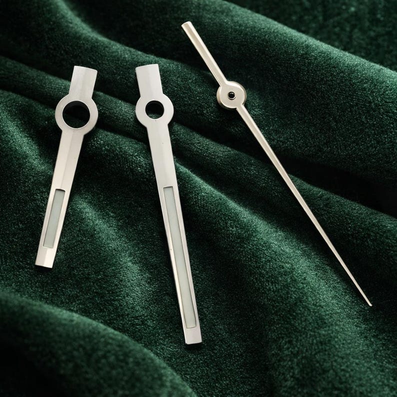 May include: Three silver watch hands on a dark green velvet surface. Two hands have a rectangular design with a circular opening and a rectangular inset. The third hand is long and thin with a circular design.