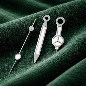 May include: Three silver watch hands with white accents are displayed against a dark green, textured background. The hands vary in design, including a thin, pointed hand with a circular detail, a rectangular hand, and a hand with a circular face and a triangular base.