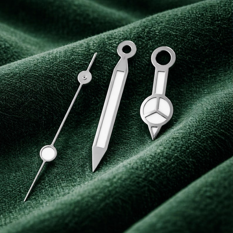 May include: Three silver watch hands with white accents are displayed against a dark green, textured background. The hands vary in design, including a thin, pointed hand with a circular detail, a rectangular hand, and a hand with a circular face and a triangular base.