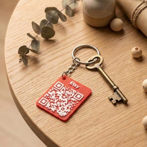 May include: A red Etsy QR code keyring with a silver key and chain. The keyring features a white QR code design and the Etsy logo. The key is bronze-coloured. The items are on a wooden table.