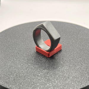 May include: A silver ring with a square face, displayed on a red square stand, on a black surface.