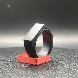 May include: A black ring with a geometric design, resting on a small, red rectangular base. The ring has a flat top and angular sides. The base is a solid, vibrant red. The ring appears to be made of metal.