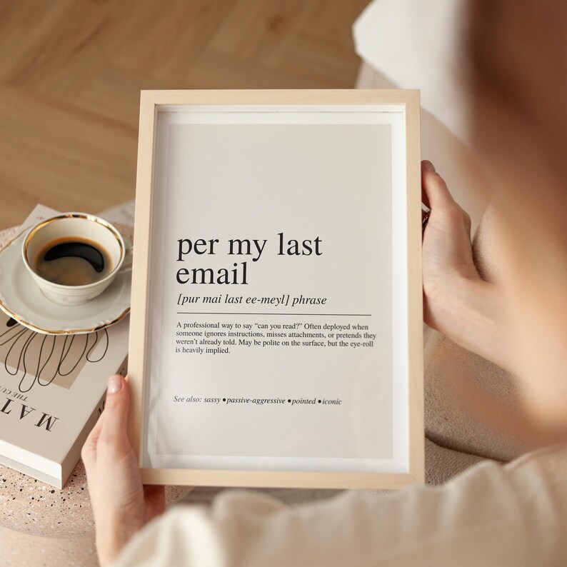 A person is holding a framed definition of "per my last email" in front of a coffee cup and a book, with a blurred background.
