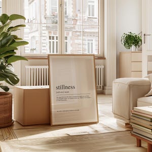 a minimalist living room with a large window, a plant, and a framed definition of stillness.