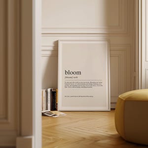 a framed definition of 'bloom' on a wall, surrounded by a bookshelf and a yellow ottoman.