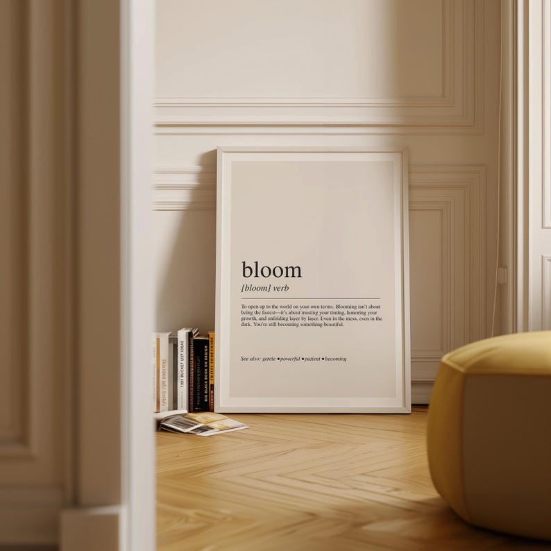 a framed definition of 'bloom' on a wall, surrounded by a bookshelf and a yellow ottoman.