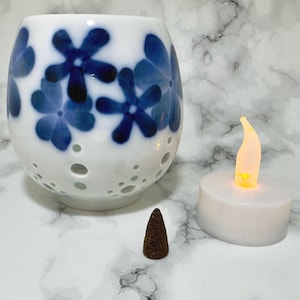 May include: A white ceramic incense burner featuring a blue floral pattern and circular cutouts at the base. Beside the burner sits a single brown cone-shaped incense piece and a small white battery-operated tea light with a glowing orange flame.