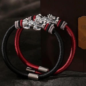 Peut inclure: Deux bracelets avec des têtes de dragon argentées et des cordons tressés. L'un est noir, l'autre rouge. Les bracelets ont des détails argentés et des fermoirs. Les têtes de dragon sont finement ouvragées.
