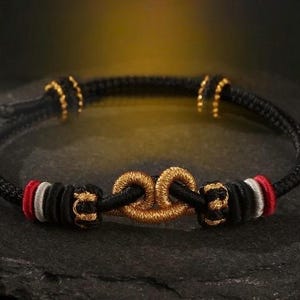 May include: Black rope bracelet with gold-colored accents and a central gold-colored knot design. The bracelet features red and white details near the knot. The bracelet is displayed on a dark, textured surface.