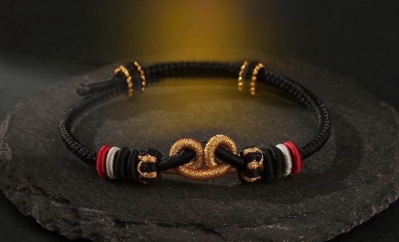 May include: Black rope bracelet with gold-colored accents and a central gold-colored knot design. The bracelet features red and white details near the knot. The bracelet is displayed on a dark, textured surface.