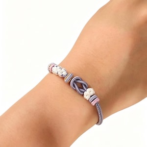 May include: A bracelet featuring a central lavender cord tied in a knot, accented with white braided knots, small pink cylindrical beads, and a gold-toned textured spacer bead, all strung on a lavender cord that wraps around the wrist.