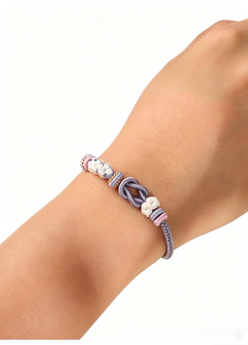 May include: A bracelet featuring a central lavender cord tied in a knot, accented with white braided knots, small pink cylindrical beads, and a gold-toned textured spacer bead, all strung on a lavender cord that wraps around the wrist.