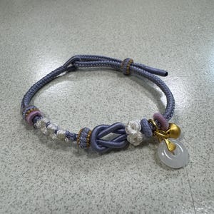 May include: A lavender cord bracelet featuring a central decorative knot, white and lavender woven accents, a small gold-toned bell, and a translucent white circular stone pendant suspended from a gold-toned metal bail on a sliding cord closure.