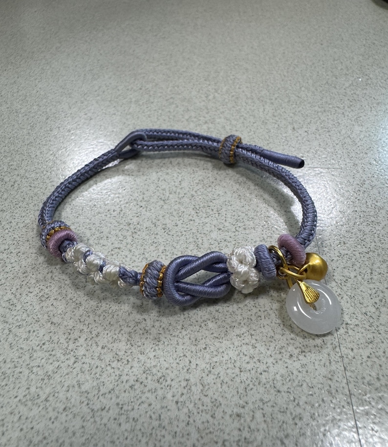 May include: A lavender cord bracelet featuring a central decorative knot, white and lavender woven accents, a small gold-toned bell, and a translucent white circular stone pendant suspended from a gold-toned metal bail on a sliding cord closure.