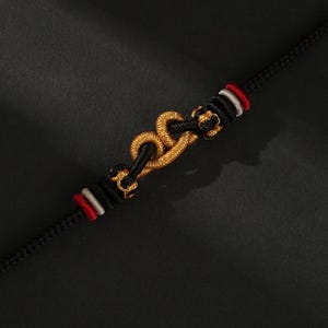 May include: A black bracelet with a gold-colored clasp. The clasp is in the shape of a decorative knot. The bracelet has red and white accents near the clasp. The bracelet is made of a woven material.