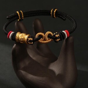 May include: A black cord bracelet featuring a central gold-colored knot ornament and a cylindrical gold-colored bead with embossed symbols. The cord is accented with small red, white, and black thread details and two gold-colored sliding adjustment beads.