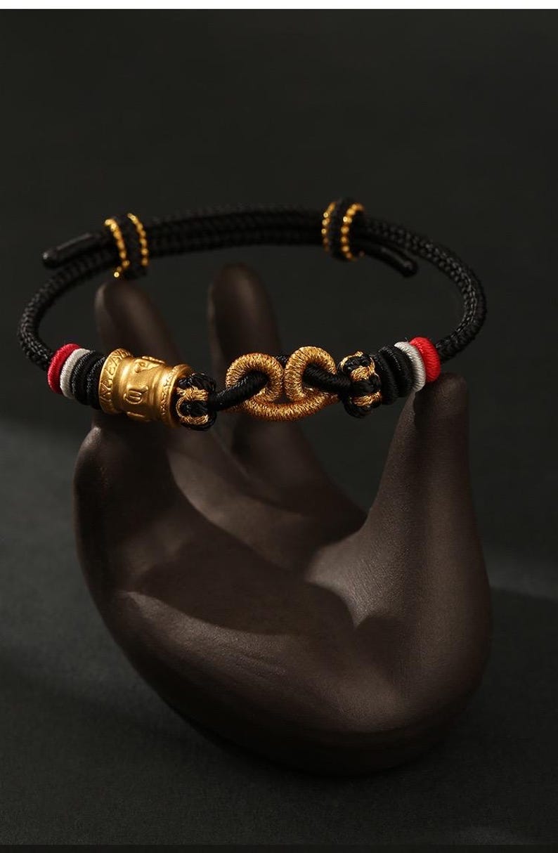 May include: A black cord bracelet featuring a central gold-colored knot ornament and a cylindrical gold-colored bead with embossed symbols. The cord is accented with small red, white, and black thread details and two gold-colored sliding adjustment beads.