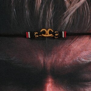 May include: A black cord bracelet featuring a gold-colored knot design at the center. The bracelet has red, white, and black accents on either side of the knot. The background is a dark, textured surface.