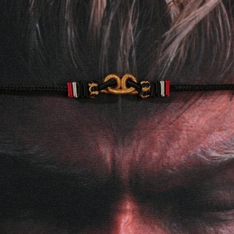 May include: A black cord bracelet featuring a gold-colored knot design at the center. The bracelet has red, white, and black accents on either side of the knot. The background is a dark, textured surface.