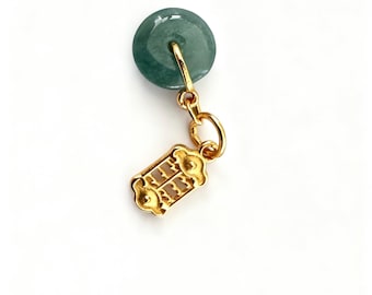 May include: A close-up of a jade green pendant with a gold-colored clasp. The pendant is round with a hole in the center. The clasp is rectangular with ornate detailing. The background is white.